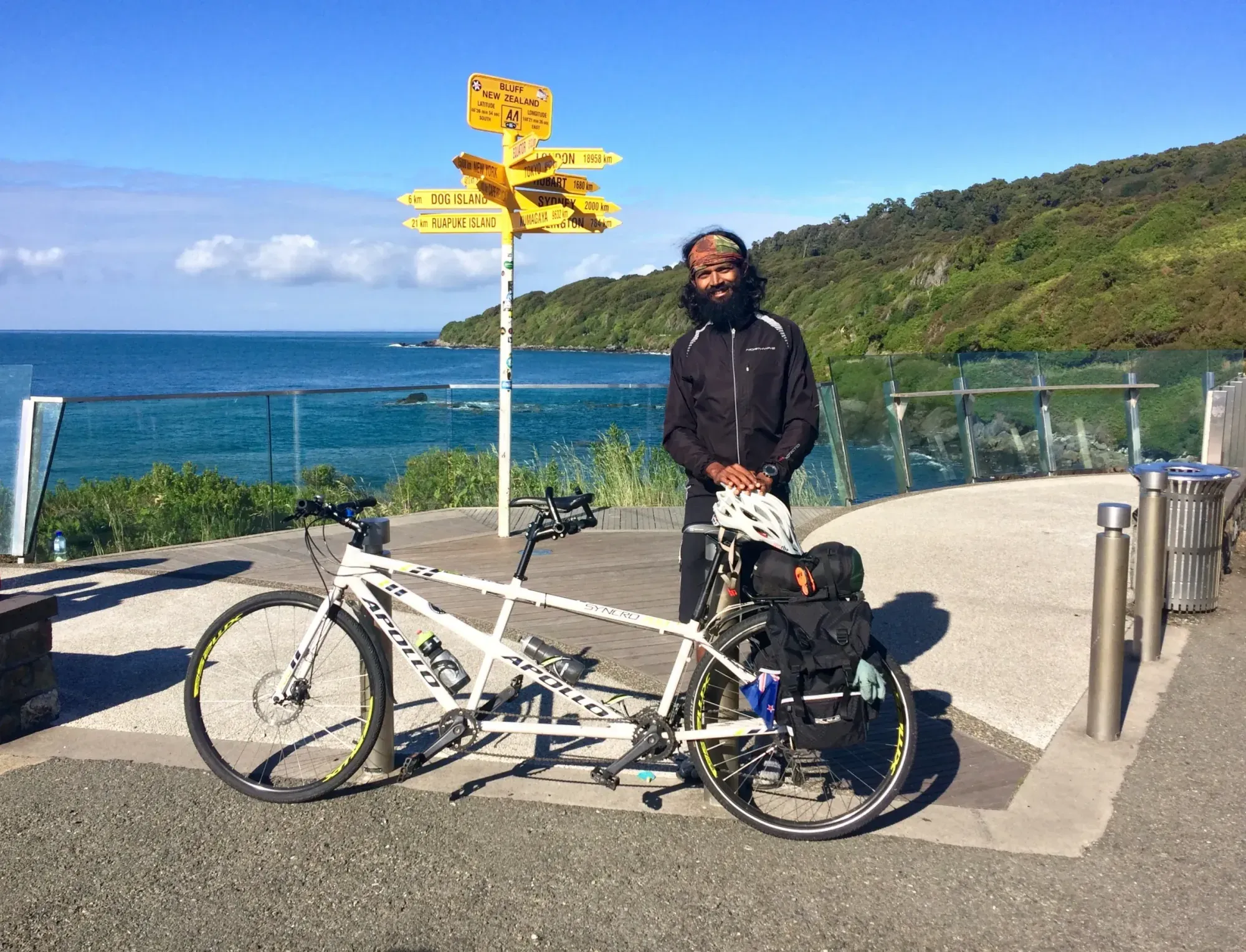 Naresh and a white tandem beside the yellow signpost at Bluff, on the coast