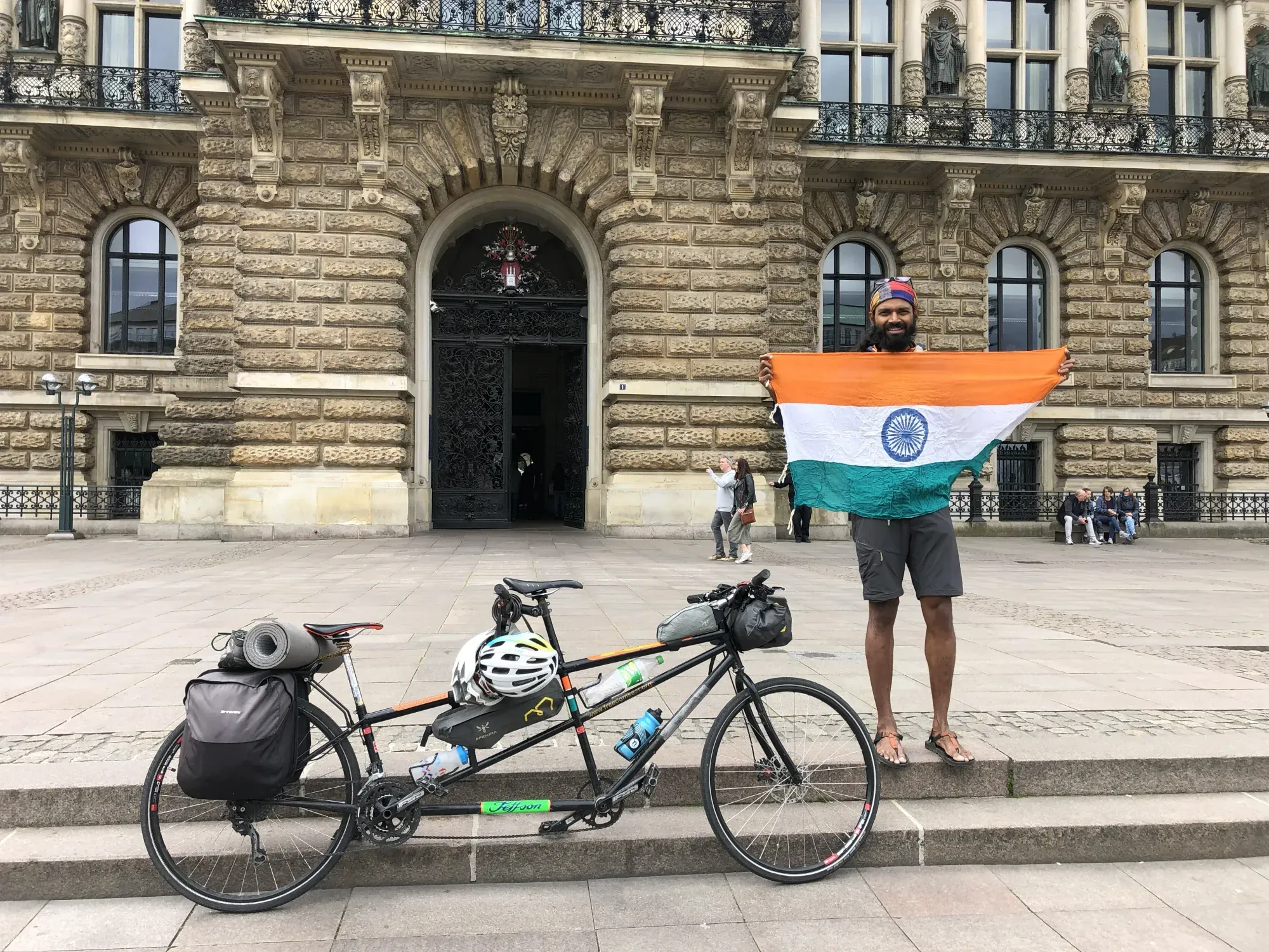 Naresh holding an Indian flag beside the tandem in front of a grand stone building