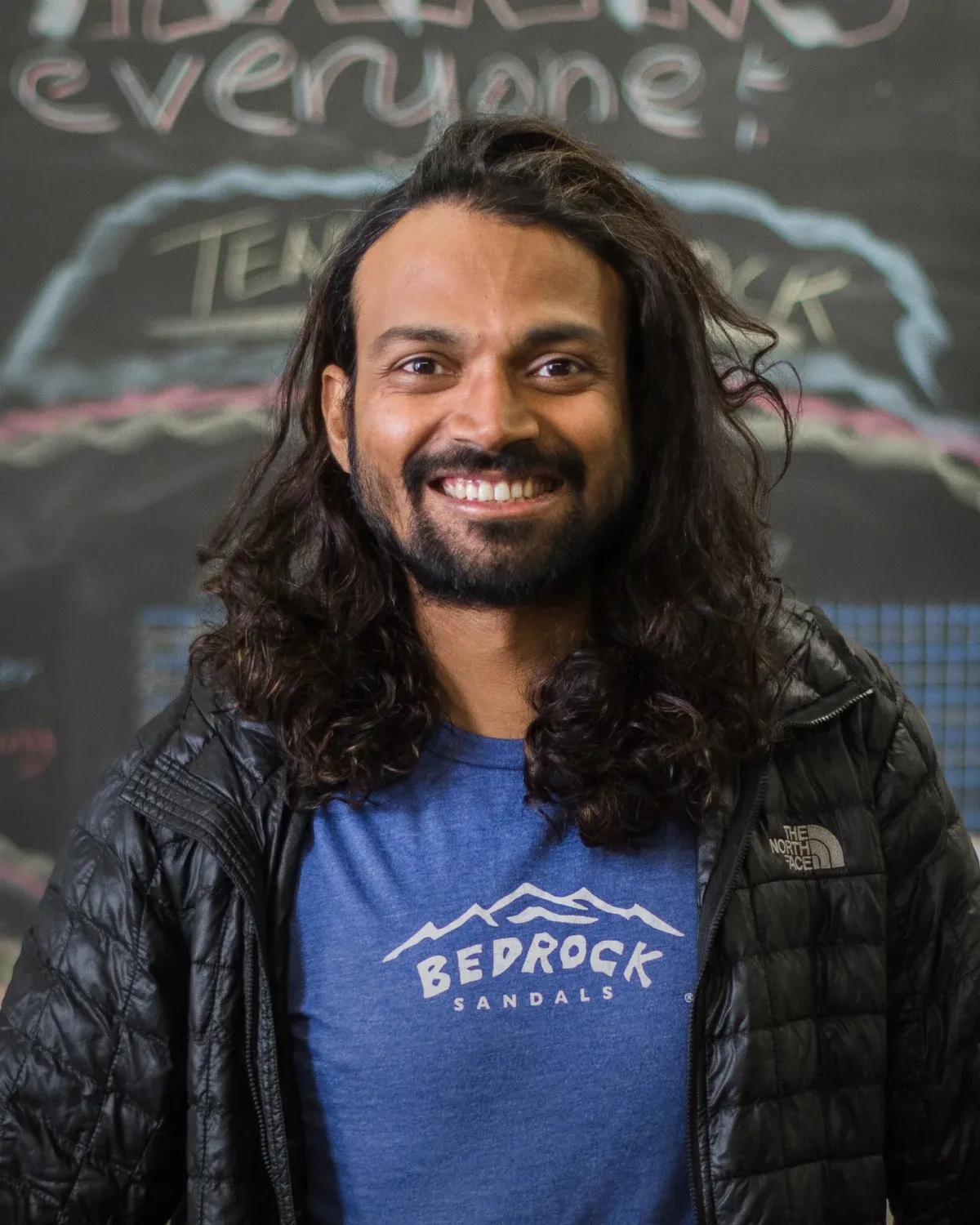 Naresh Kumar smiling, with long curly hair, in a black North Face jacket and a Bedrock Sandals T-shirt